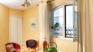 A room with a dining table, chairs, a window with curtains, a green kettle, a bottle, and a loaf of bread.
