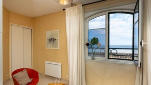 A room with a red chair, a small wooden table with a plate and glasses, a radiator, a window with a view of the sea, and a painting on the wall.