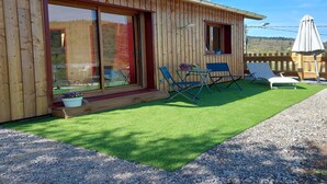A wooden cabin with a sliding glass door, a small patio with two blue chairs and a table, and a gravel path leading to the door.