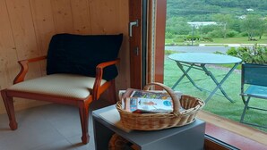 A wooden chair with a cushion, a wicker basket with magazines, and a view of a green landscape through a glass door.