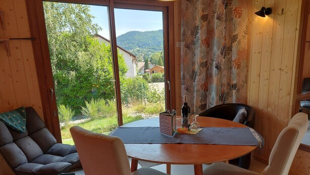 A dining area with a table and chairs, a view of greenery and buildings outside, and a wooden interior.