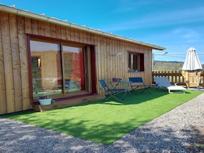 Een houten gebouw met een schuifdeur van glas, een klein terras met kunstgras en twee blauwe stoelen.