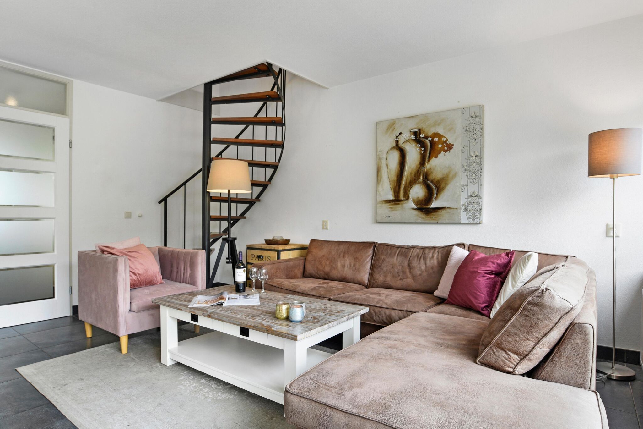 Un salon moderne avec un escalier en colimaçon, un canapé beige, un fauteuil rose et une table basse.