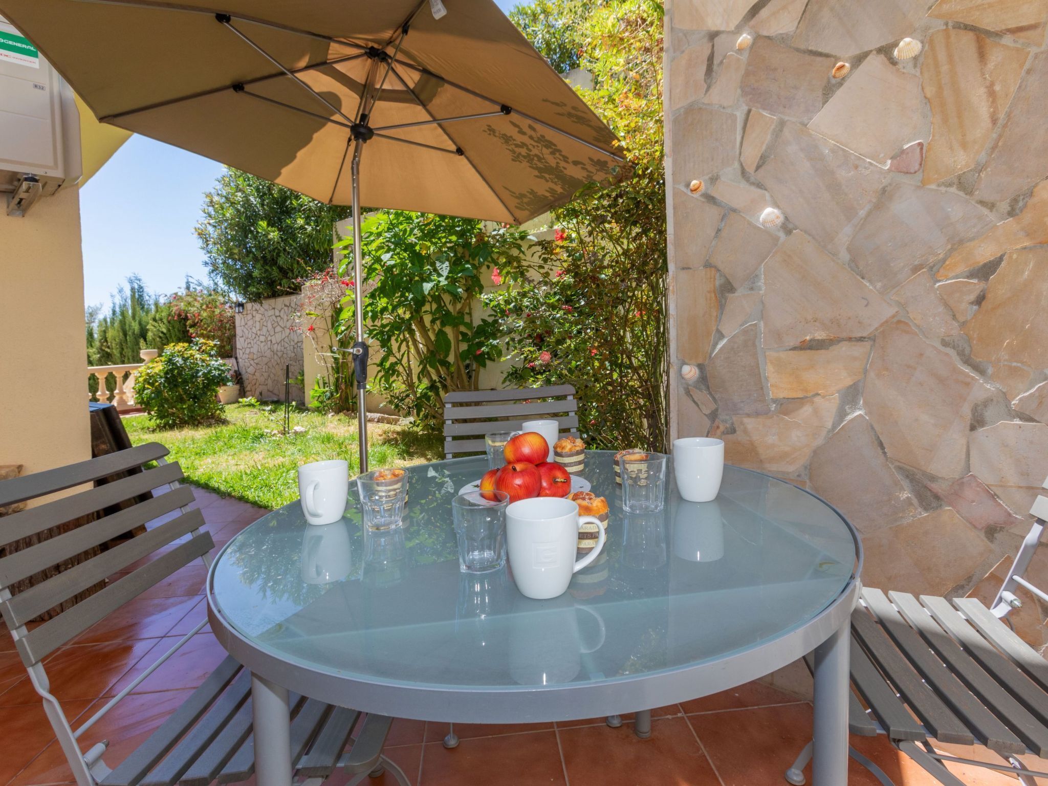 Une table en verre surmontée d’un parasol beige, entourée de chaises et adossée à un mur en pierre.