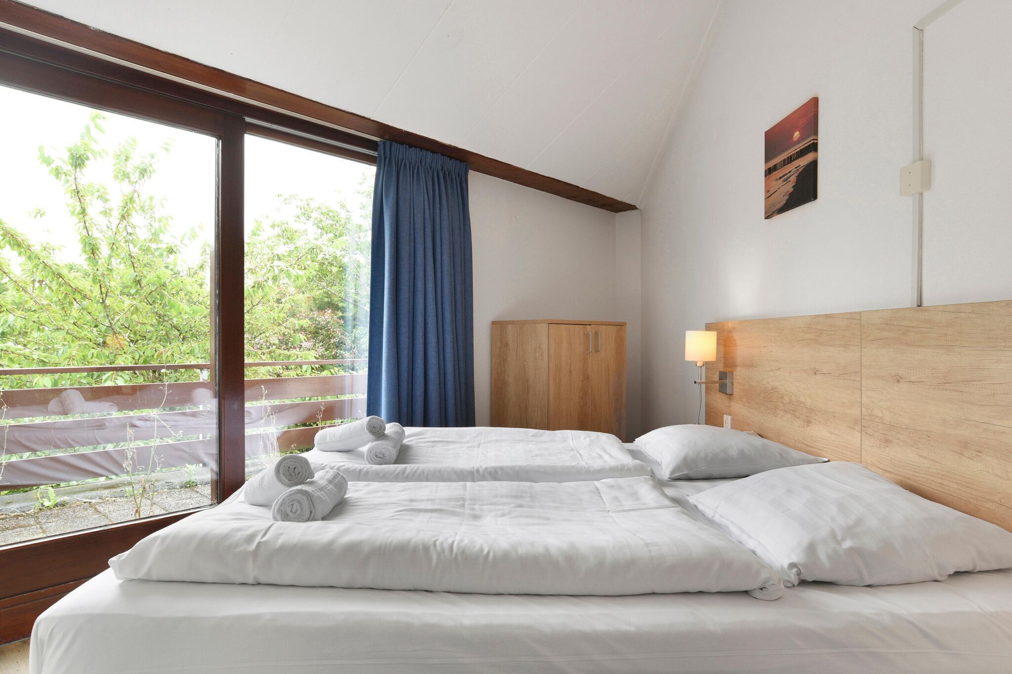 A hotel room with two beds, a wooden headboard, a wardrobe, and a window with a view of greenery.