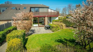A modern two-story house with a covered patio, a lawn, and flowering trees. - Holiday Home by Grevelingenmeer & Brouwersdam (Brouwershaven)