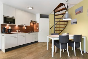 Et moderne køkken med hvide skabe, en træbordplade og en indbygget mikroovn. Der er et spiseområde med et hvidt bord og sorte stole. En spiraltrappe med gelænder i træ fører op til et andet niveau.