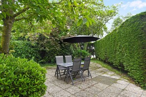 Un patio exterior con una mesa y sillas, rodeado de abundante follaje y un seto.