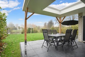 Une terrasse extérieure avec une table et des chaises, un espace abrité et une vue sur une pelouse et des arbres.