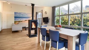 A modern living room with a wooden floor, a fireplace, a dining table with blue chairs, a sofa, and a large window with a view of trees and a beach.