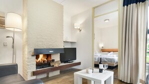 A modern hotel room with a fireplace, a flat-screen TV, a bed, a bedside table with wine glasses, and a large mirror.