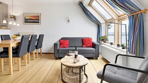 Een moderne woonkamer met een grijze bank, een eettafel met stoelen, een ronde salontafel, en uitzicht op zee door grote ramen.