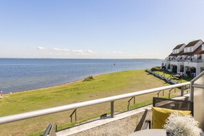 A coastal hotel with a balcony overlooking the sea, a grassy area, and a building with multiple windows.