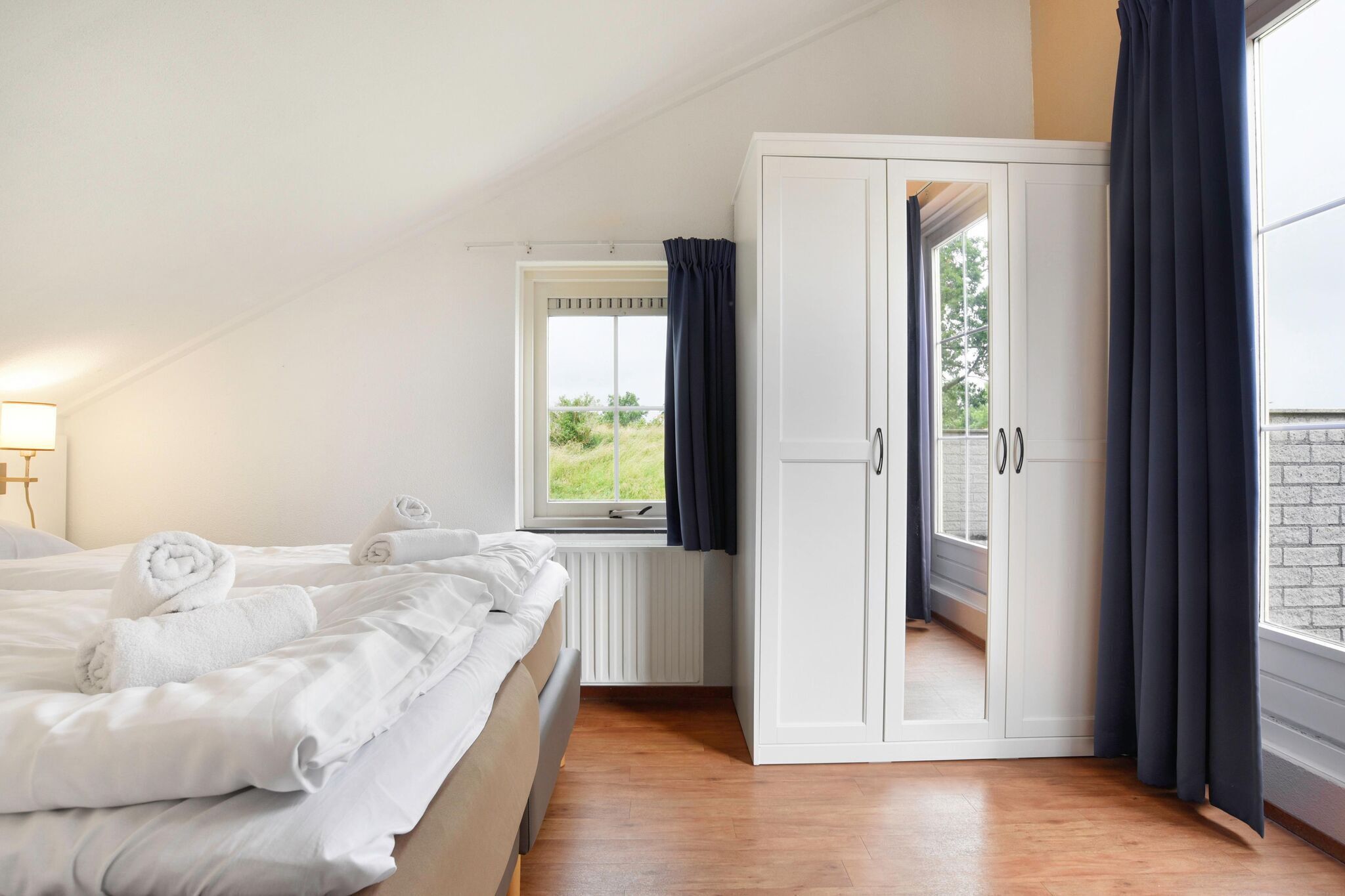 A bedroom with a bed, a window with curtains, a wardrobe, and a view of greenery outside.