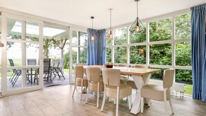 Een eethoek met een grote tafel en stoelen, grote ramen met blauwe gordijnen en uitzicht op groen buiten.