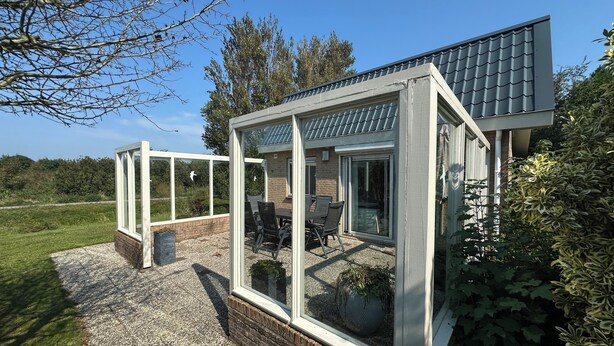 Een modern huis met een glazen zonnewerkplaats, een terras met tuinmeubilair en uitzicht op de omliggende begroeiing.