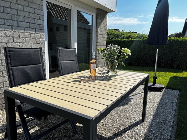 An outdoor patio with a wooden table, two chairs, a vase with flowers, and a bottle of wine.