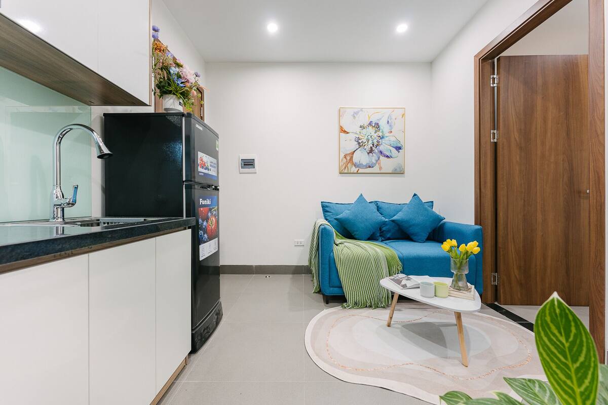 Un espai habitable reduït amb una cuineta, un sofà blau, una taula de cafè petita i una porta.
