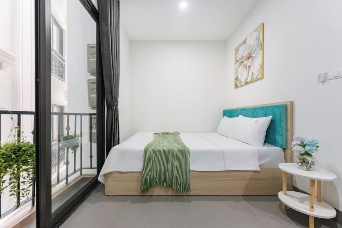 Un dormitorio con cama, una mesita de noche con un florero, un cuadro y un balcón con plantas.