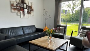 Un salon moderne avec un canapé en cuir noir, une table basse en bois ornée d’un vase de fleurs et de verres à vin, et une vue sur une pelouse verdoyante à travers de grandes fenêtres.