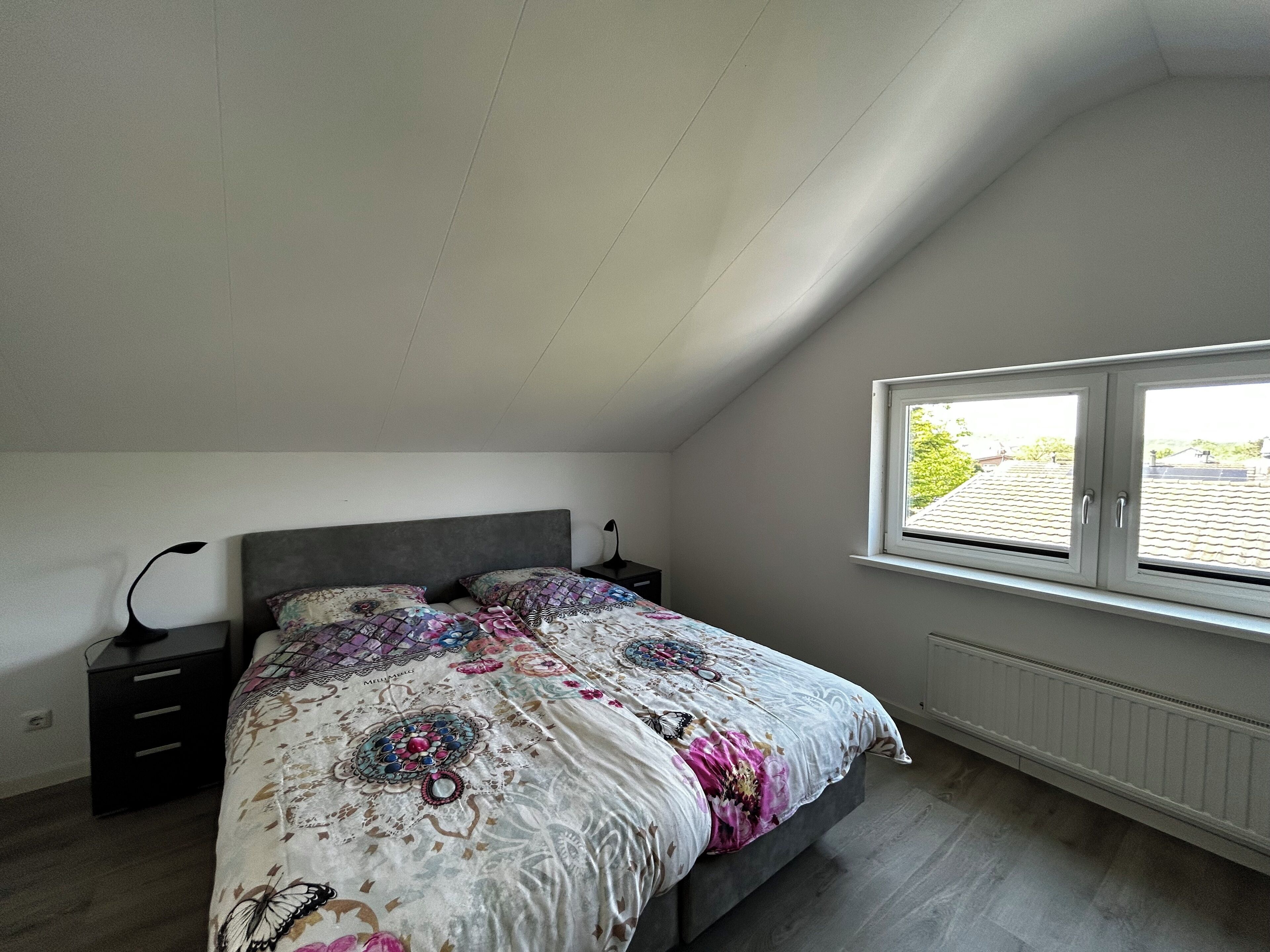 Un dormitorio con cama, mesita de noche, ventana y radiador.