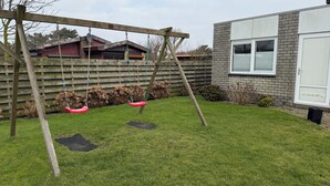 A wooden swing set in a backyard with a grassy lawn, a brick house, and a wooden fence.
