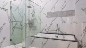 Un baño moderno con ducha envuelta en vidrio, un lavamanos blanco sobre encimera negra y paredes con estampado de mármol.