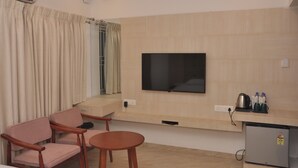 A modern hotel room with a flat-screen TV mounted on the wall, a small kitchenette with a refrigerator, a wooden table and chairs, and a window with curtains. - Green Royale Resorts (Tarangambadi)