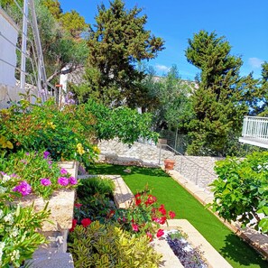 Un jardin bien entretenu, agrémenté de diverses plantes et fleurs, un mur de pierre et un ciel d’un bleu limpide.