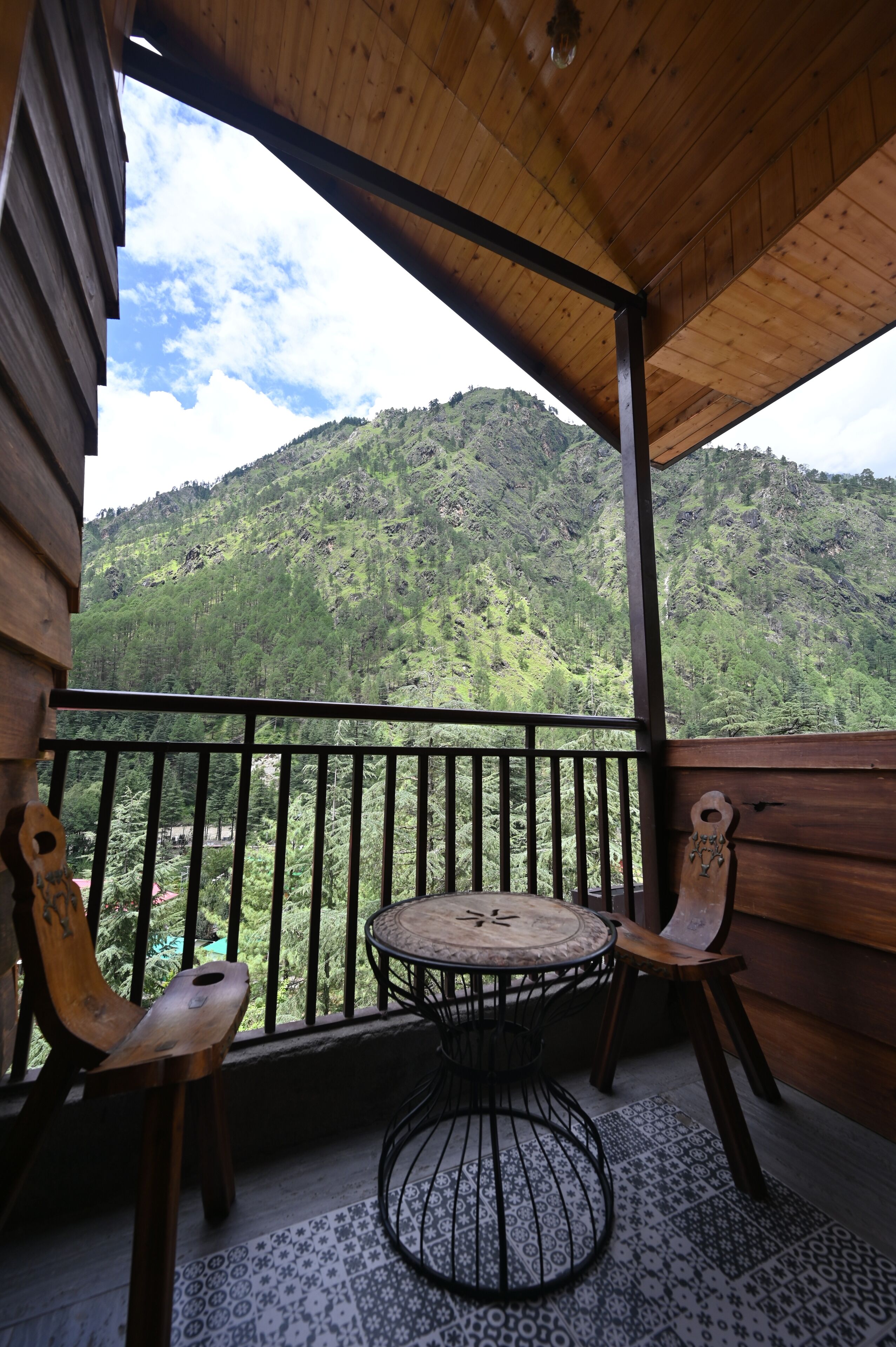 A wooden balcony with a round table and two chairs, overlooking a mountainous landscape.