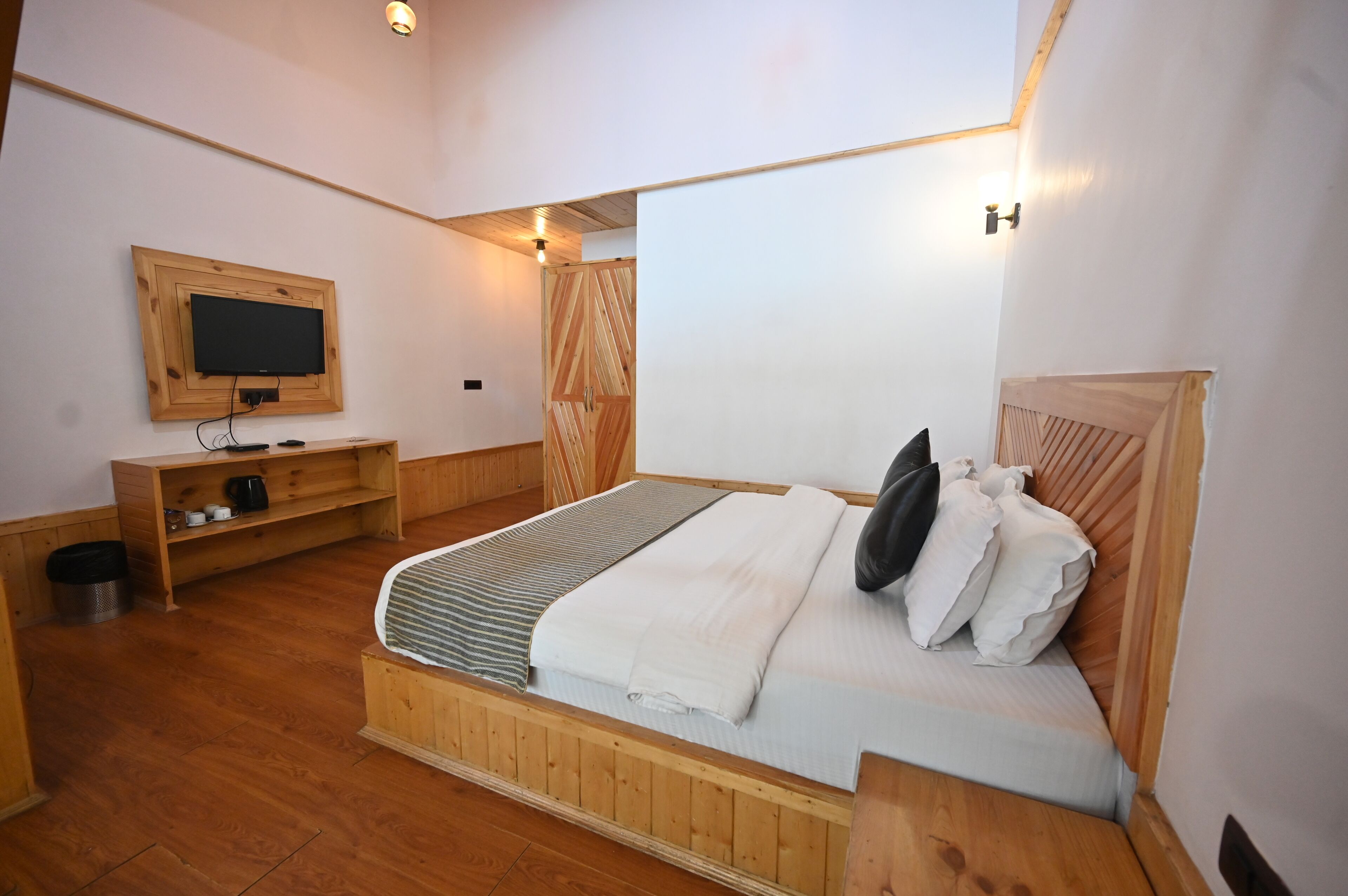 Un dormitorio con una cama de madera, un televisor montado en la pared y un pequeño estante de madera con un cubo de basura.