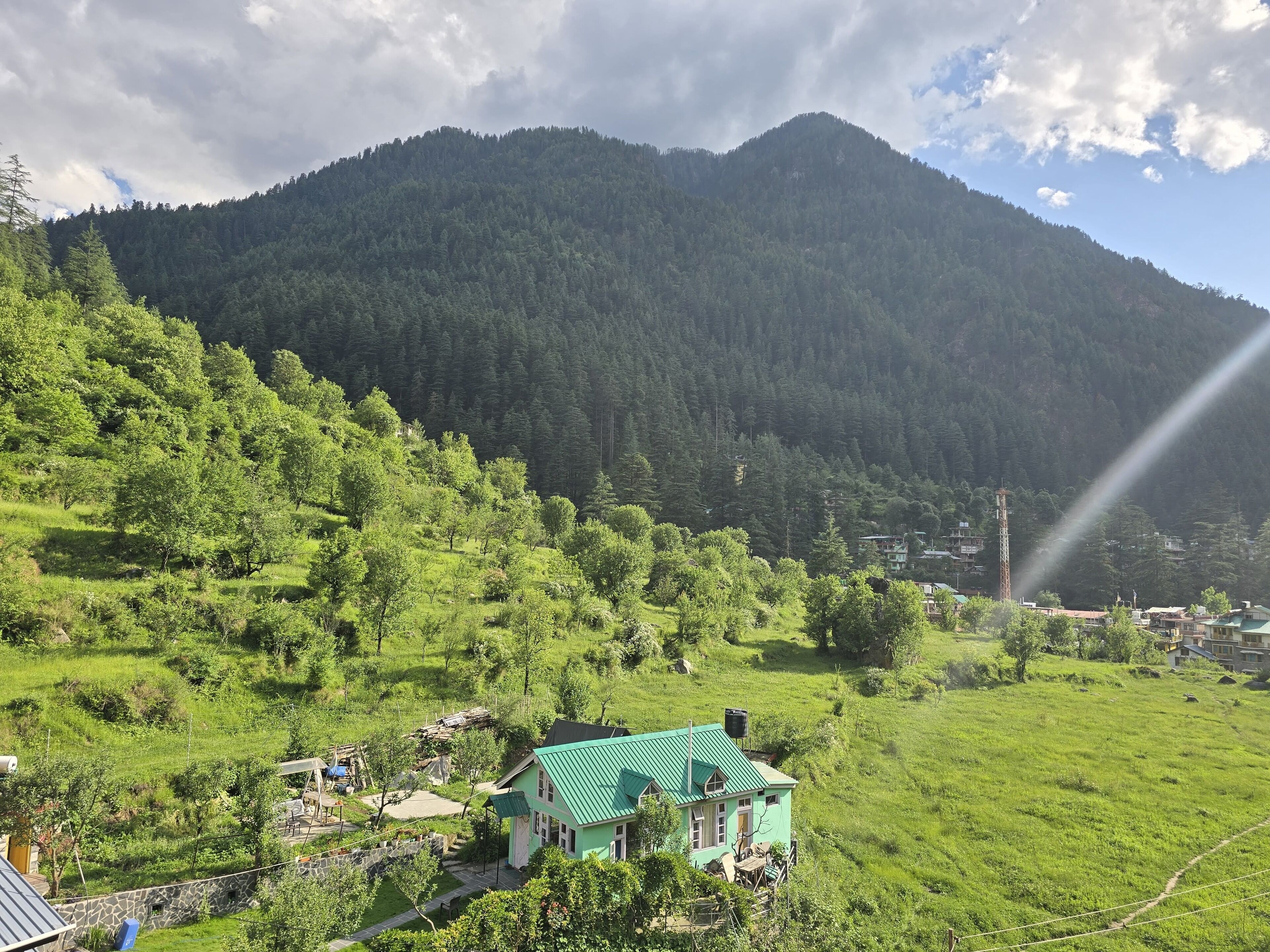 A green house with a sloped roof, surrounded by lush greenery and a mountain backdrop.