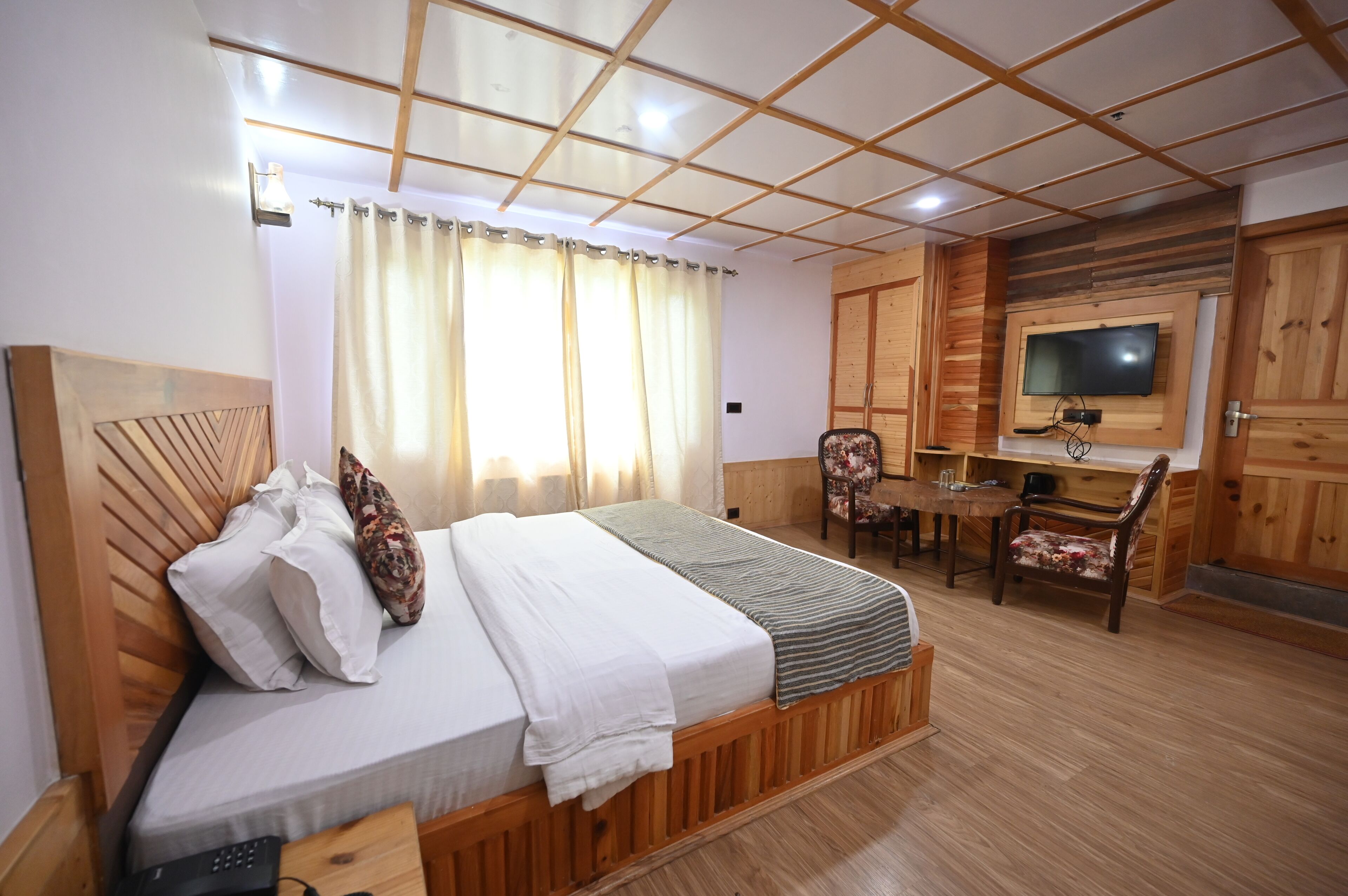 A bedroom with a wooden headboard, a bed with white linens, a bedside table with a remote control, a small desk with a chair, and a television mounted on the wall.
