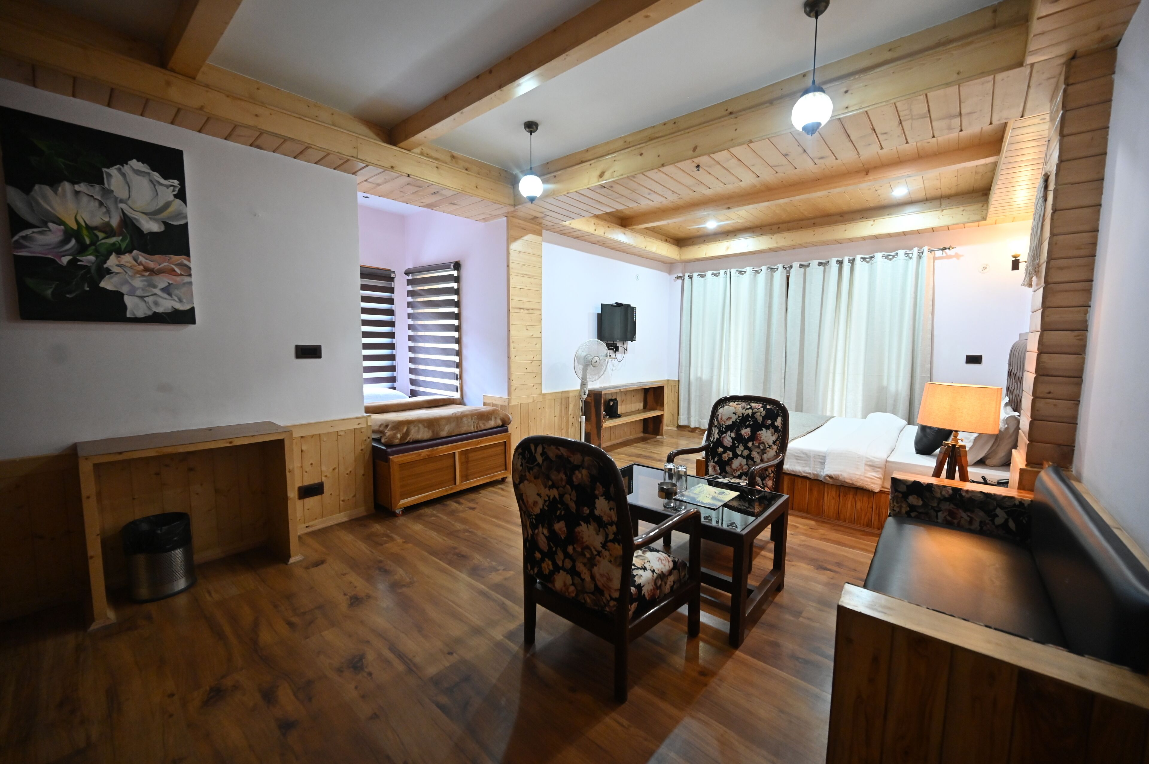 A cozy room with wooden floors, a sofa, a dining table with chairs, a bed with a nightstand, and a ceiling fan.