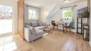 Un salon moderne avec un canapé d’angle gris, une table à manger avec des chaises, un téléviseur fixé au mur et de grandes fenêtres laissant entrer la lumière naturelle.