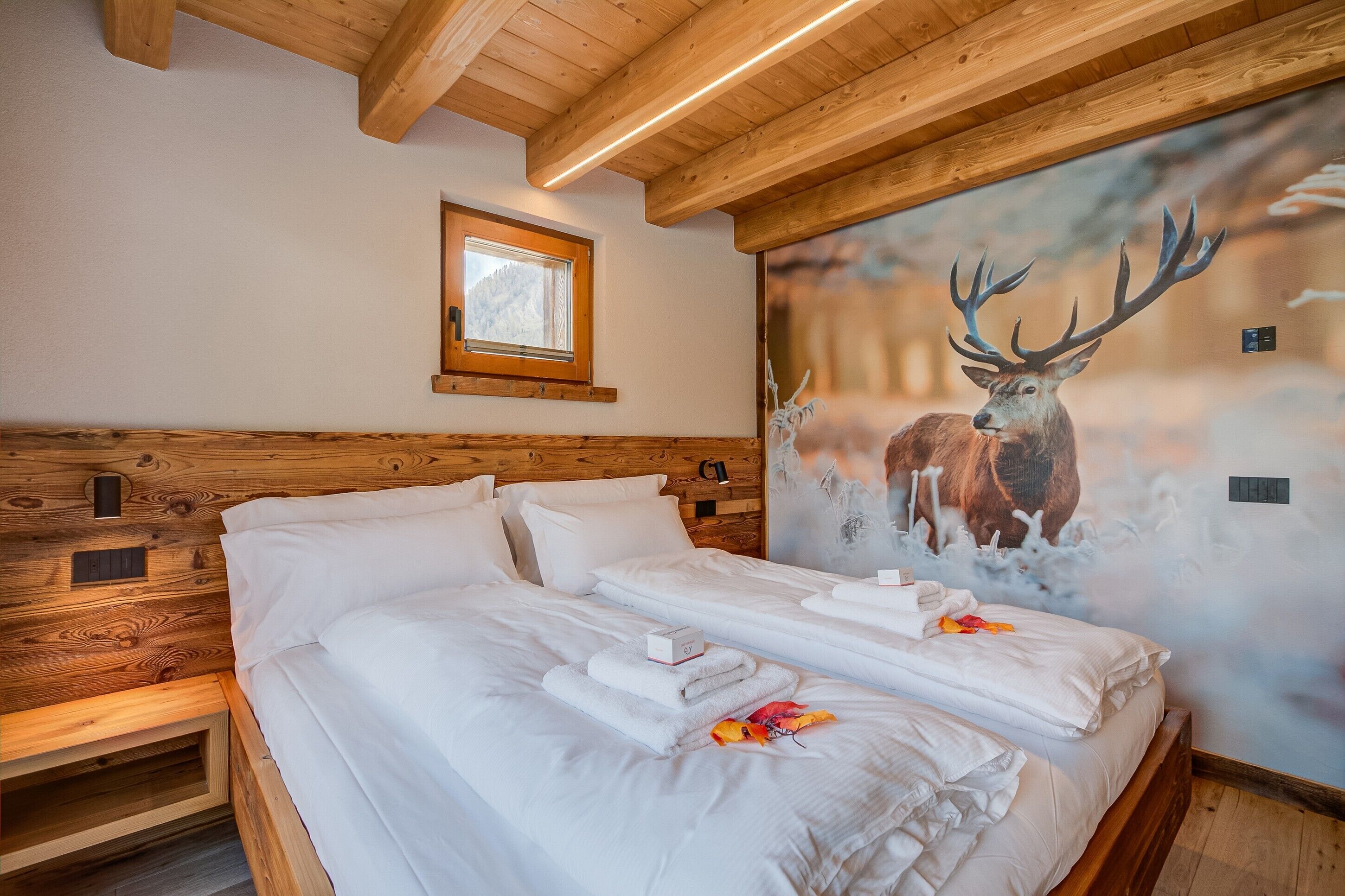 A room with wooden beams, a bed with white linens, a window, and a deer mural.