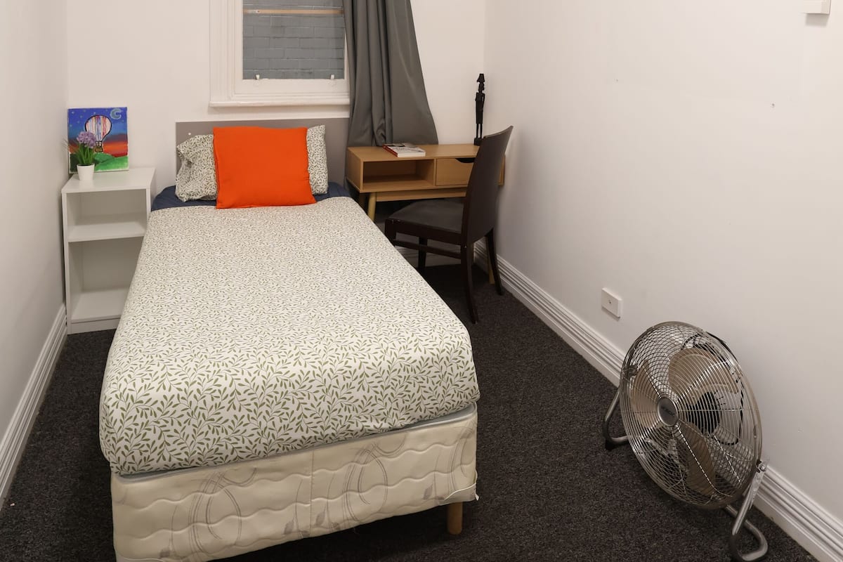 A bedroom with a bed, a desk, a chair, a standing fan, and a window with curtains.
