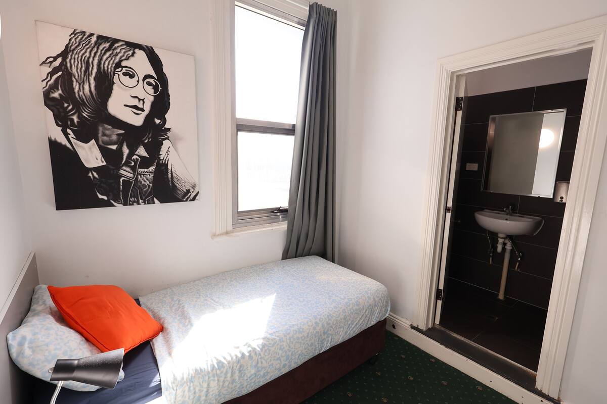 A small bedroom with a single bed, a window with curtains, a bathroom with a sink, and a wall-mounted mirror.