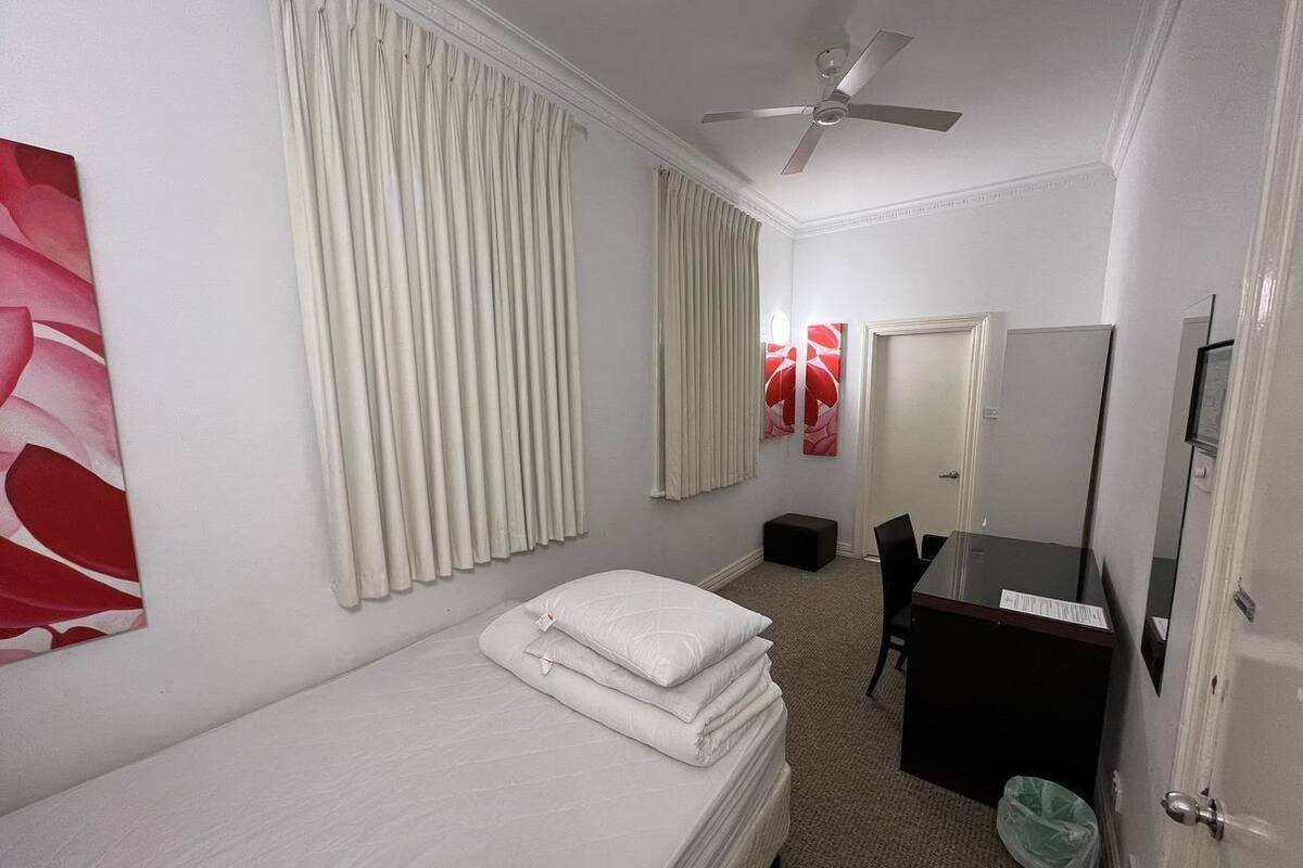 A hotel room with a single bed, a desk with a chair, a mirror, and a ceiling fan.