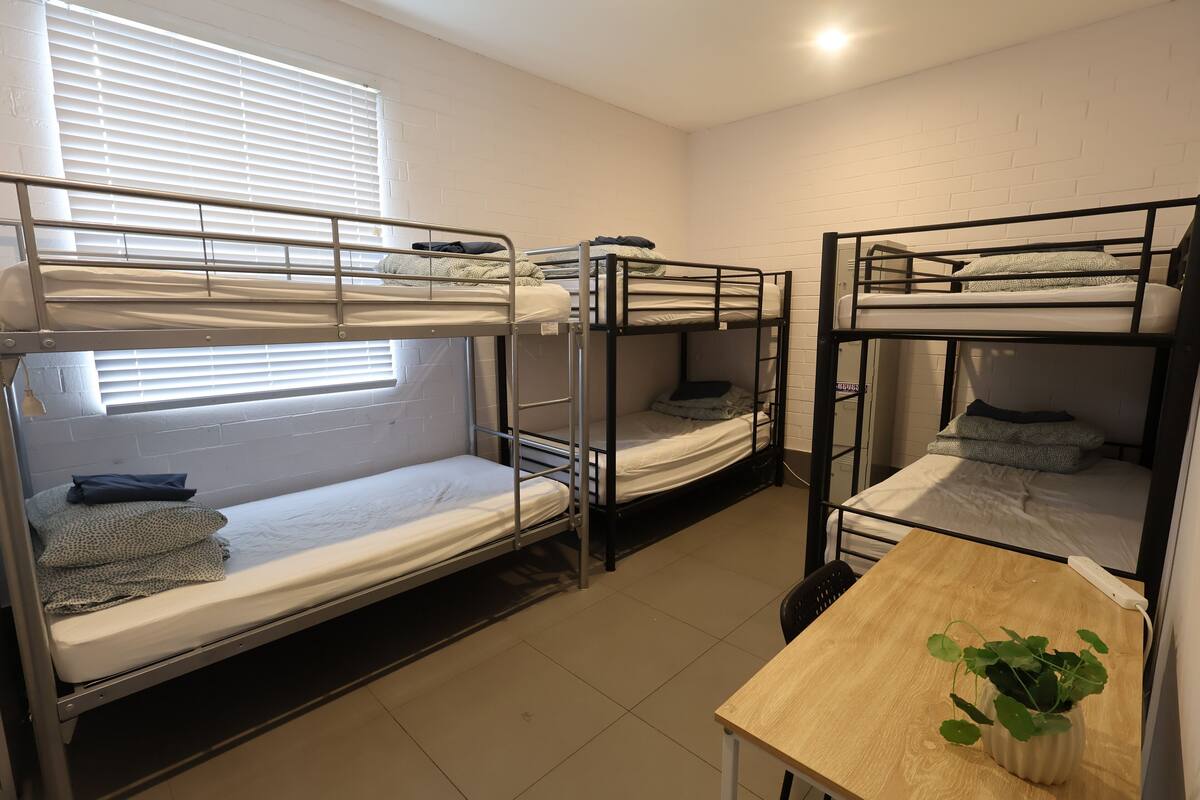 A dormitory room with bunk beds, a small table, and a window with blinds.