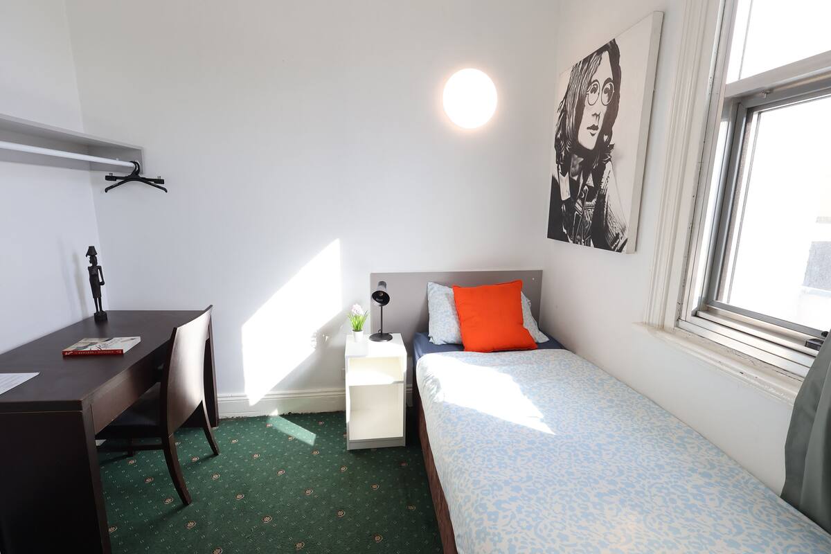 A small, neatly arranged bedroom with a desk, a bed with patterned bedding, a bedside table with a lamp, and a wall-mounted artwork.