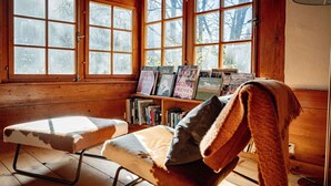 A cozy cabin interior with a wooden bench, a bookshelf with magazines, and large windows offering a view of trees.