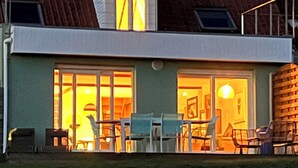 A two-story house with a large glass door and windows, a visible outdoor seating area with chairs, and a grassy yard.