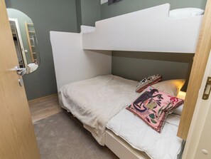 A bunk bed with a white headboard and a single bed below. There is a mirror on the wall to the left, a door on the left, and a small rug on the floor.