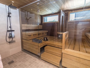 A sauna room with wooden benches, a glass partition, and a shower area with a handheld showerhead. - Casa krogerus ruka by Interhome (Kuusamo)