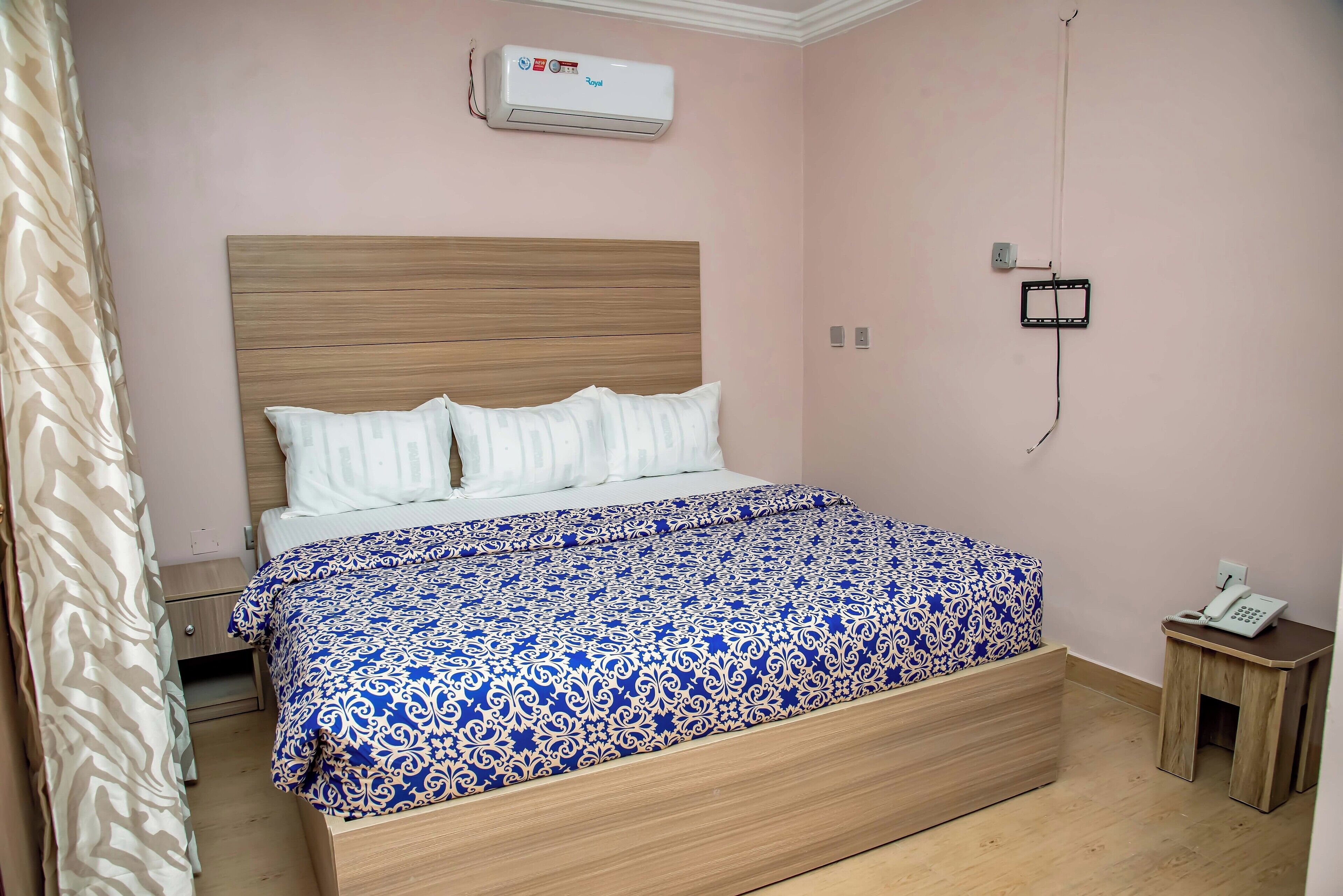 A bedroom with a wooden bed, a patterned bedspread, a bedside table with a phone, and an air conditioning unit on the wall.
