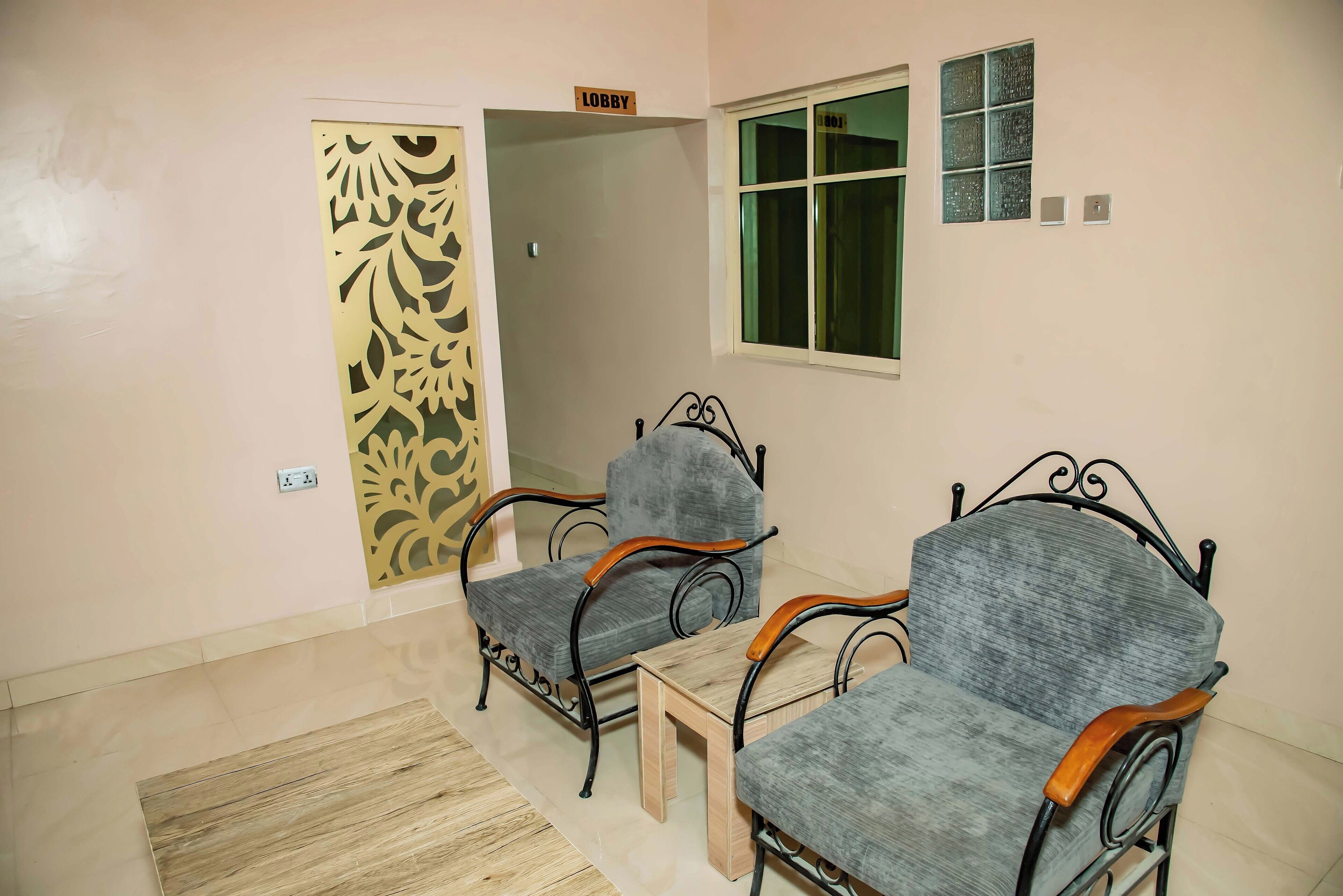 A room with two grey upholstered chairs with wooden armrests, a small wooden table, a decorative panel on the door, a window with a green view, and a sign indicating the room is the lobby.