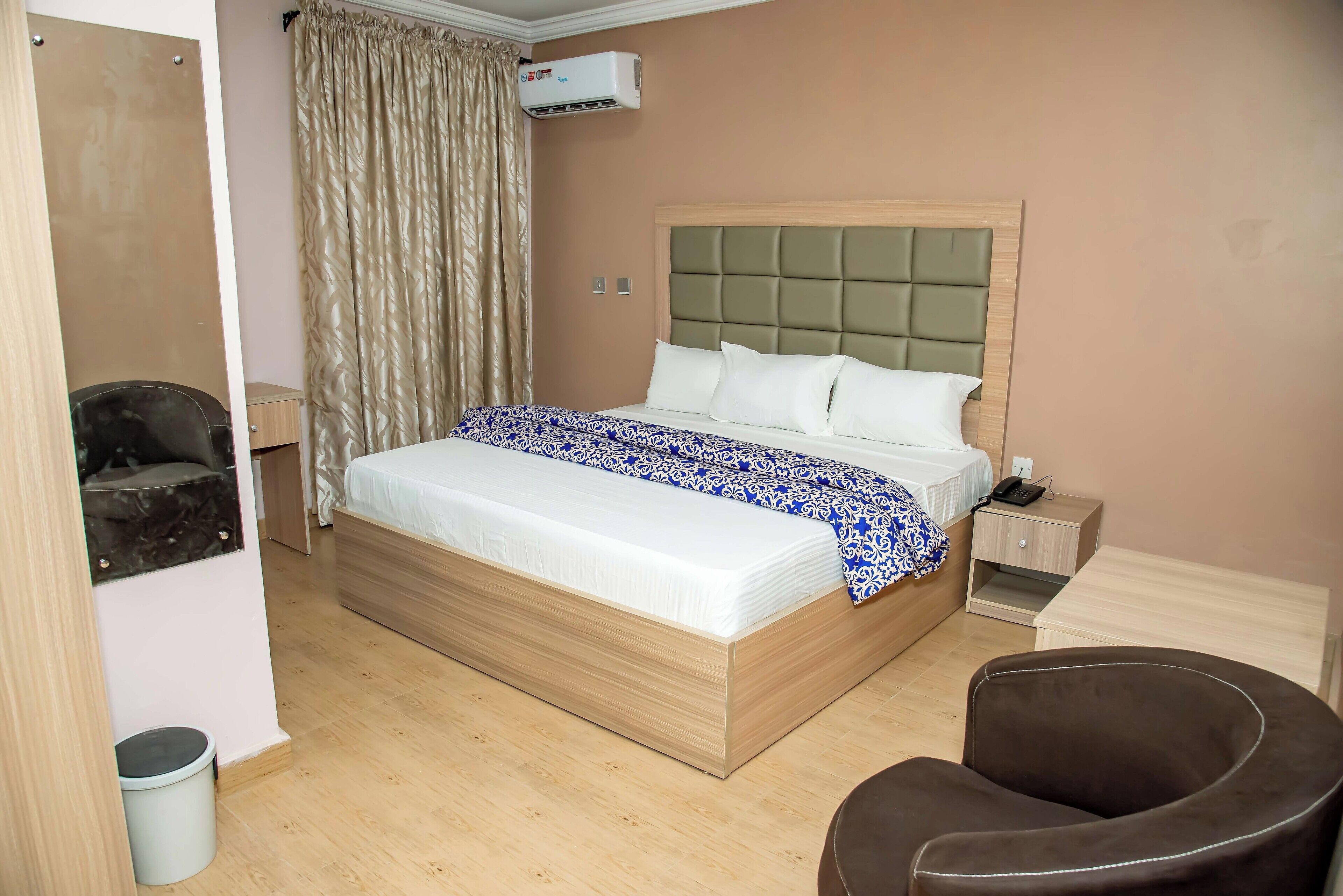 A hotel room with a large bed, a bedside table, a chair, and an air conditioning unit.