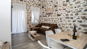 Una sala de estar acogedora con una pared de piedra, un sofá marrón, una mesa de centro de madera y una mesa de comedor con copas de vino y una botella.