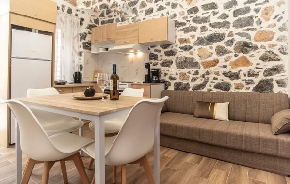 A cozy kitchen with a stone wall, a wooden dining table, white chairs, and a brown sofa. - City Stone Houses Kalymnos  (Kalymnos)